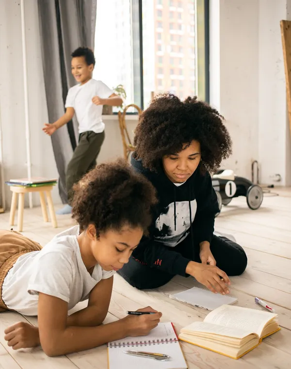 Une maman accompagne ses enfants dans leurs devoirs, assise par terre avec eux