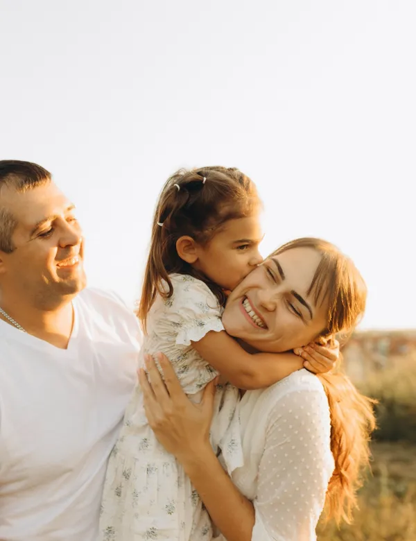 Une petite fille embrasse sa maman sur la joue, sous le regard amusé de son papa
